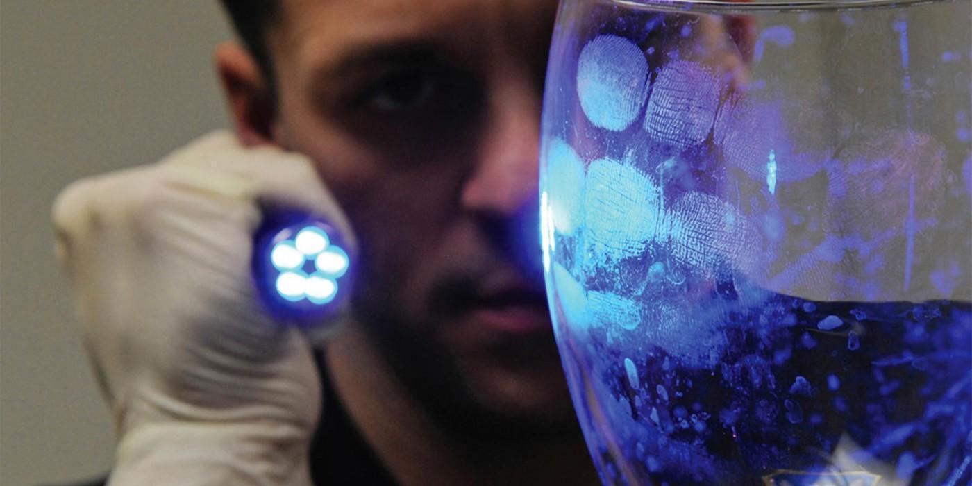 A forensic investigator wearing gloves uses a UV flashlight to reveal fingerprints on a glass surface, illustrating crime scene investigation techniques studied in a Criminology BSc degree course.