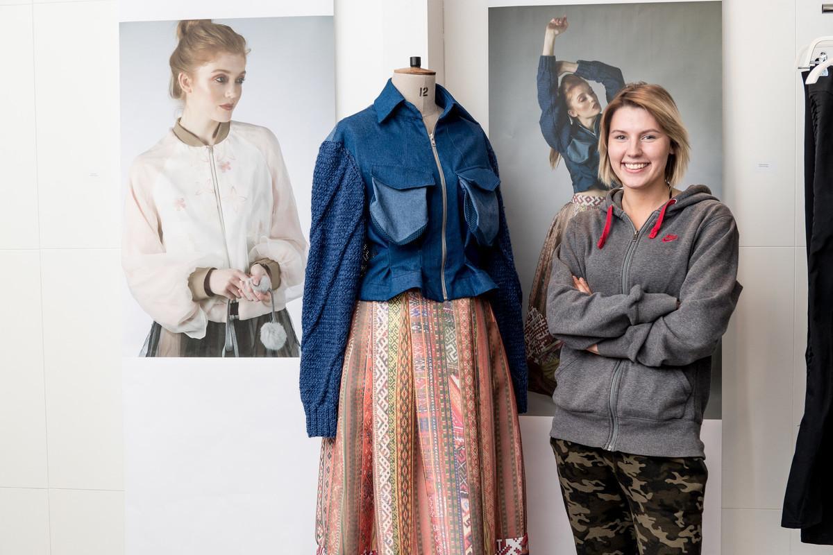 Fashion design student standing beside a mannequin showcasing their creative garment, featuring a blue textured jacket and patterned skirt, with fashion photography displayed in the background—illustrating innovation and craftsmanship in a Fashion Design BA (Hons) course.