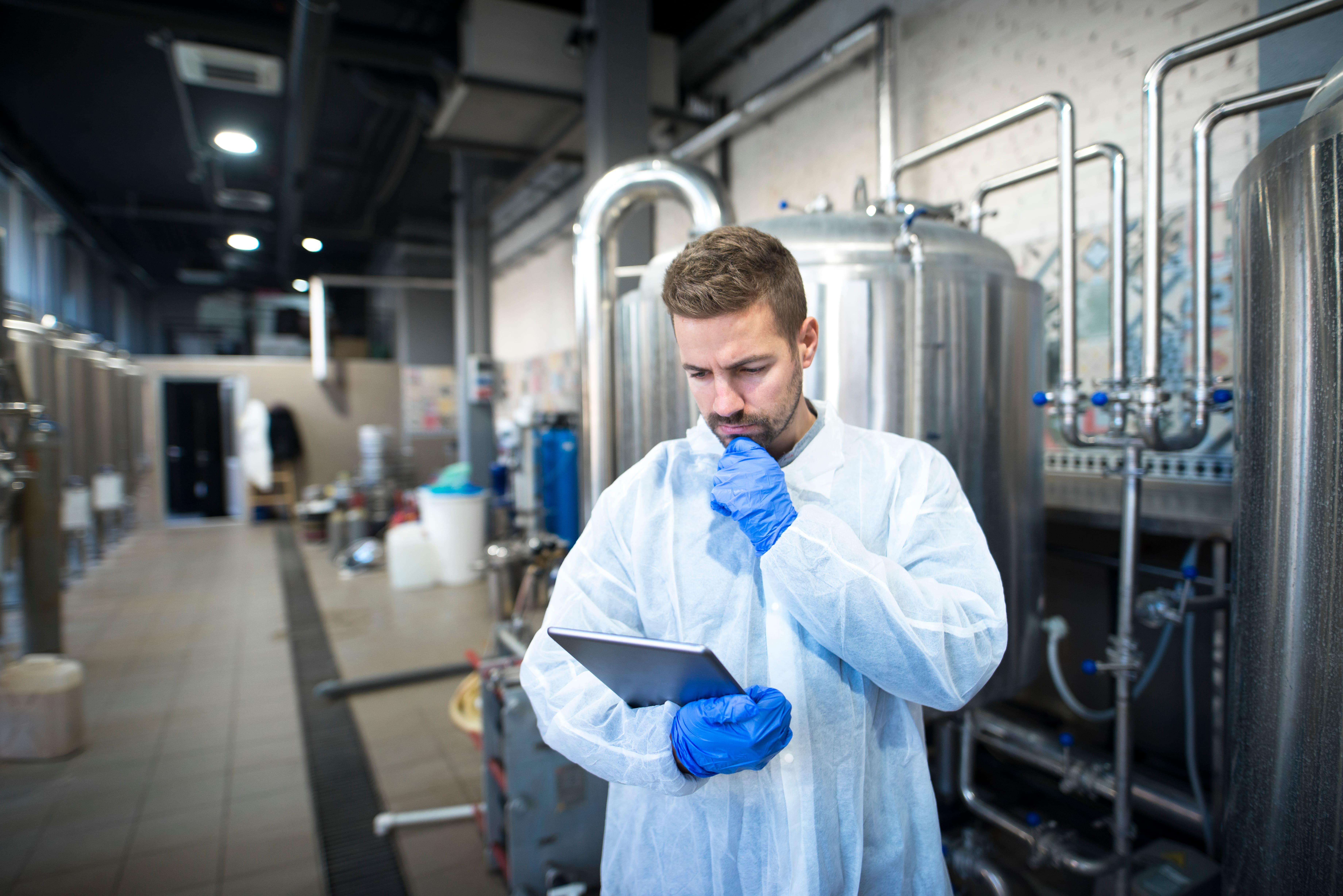 Chemical engineering Beng Hons student conducting process analysis on a digital tablet within an industrial laboratory setting, surrounded by stainless steel reactors, storage tanks, and process piping.