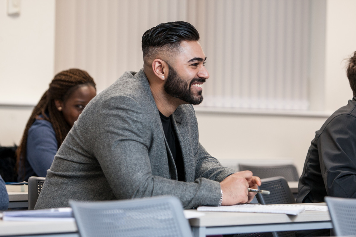 A male student smiling while sitting in a lecture room, taking notes with pen and notebook. Two more students are sitting in other rows of grey coloured seats.