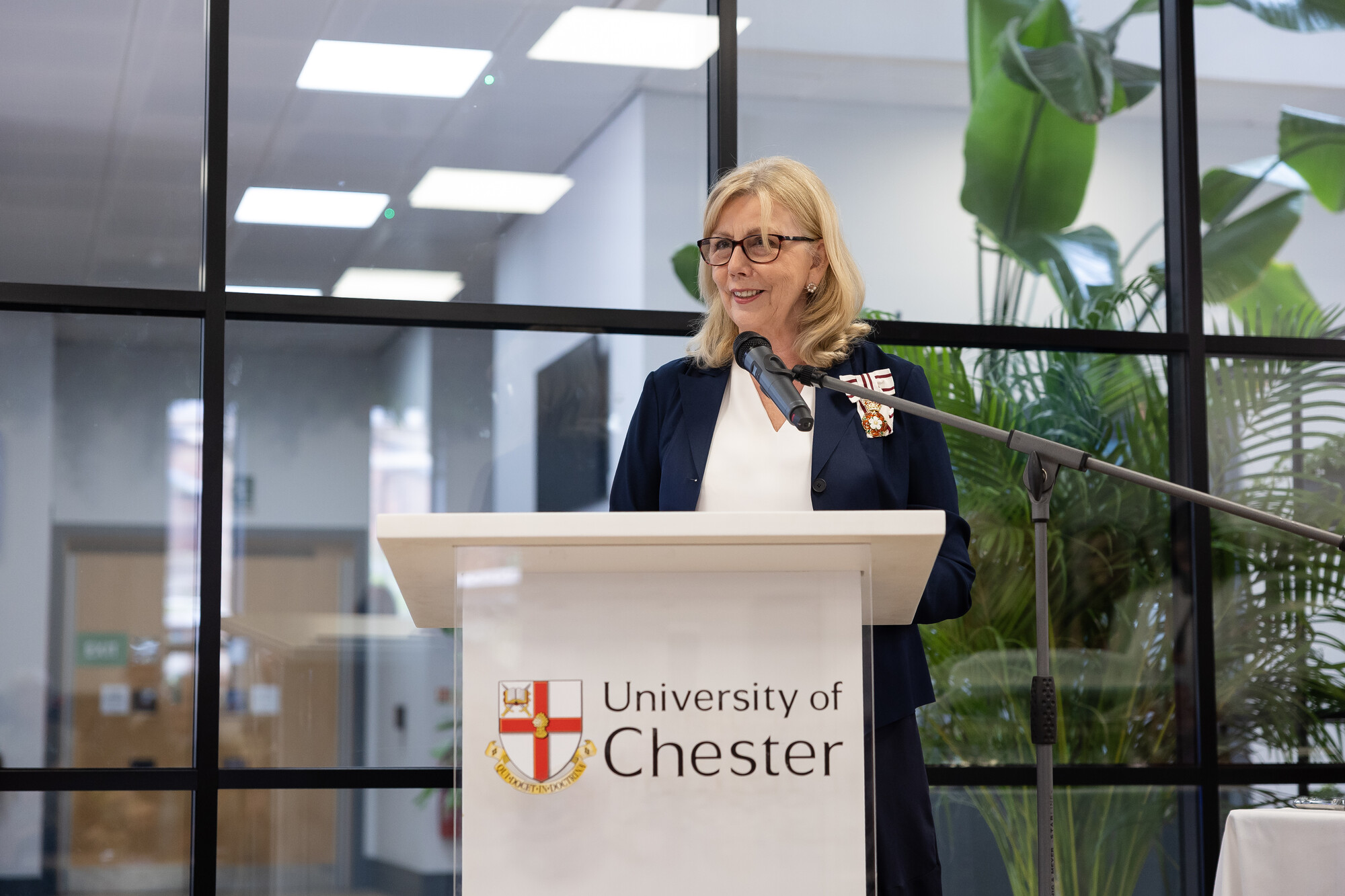 Lady Redmond MBE at the opening of Binks 2026, standing behind a university of Chester plinth, with large, bright rooms and indoor plants in the background