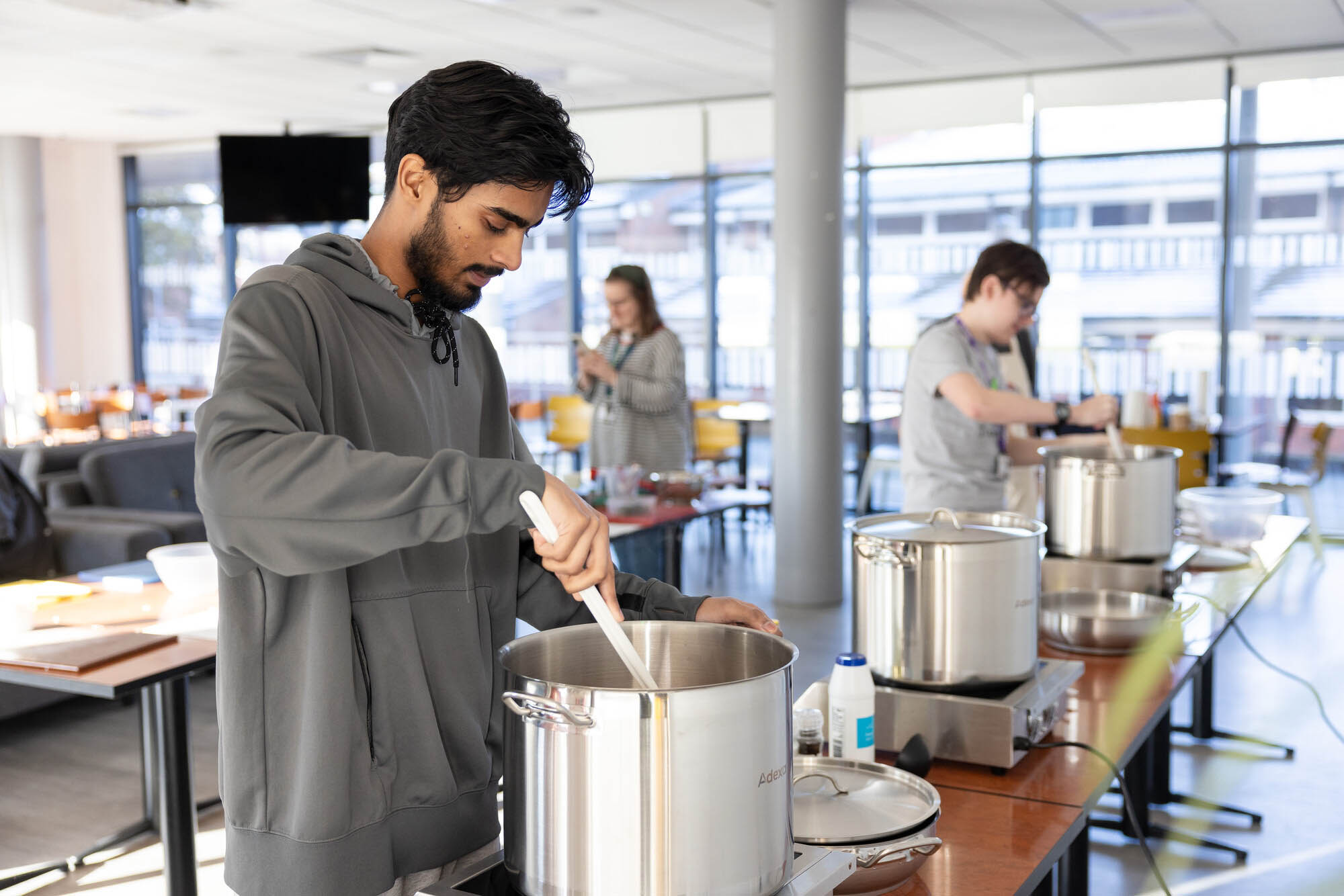 Person stirring a large stainless steel pot with a white spatula during a communal cooking event in a bright, modern kitchen setting.