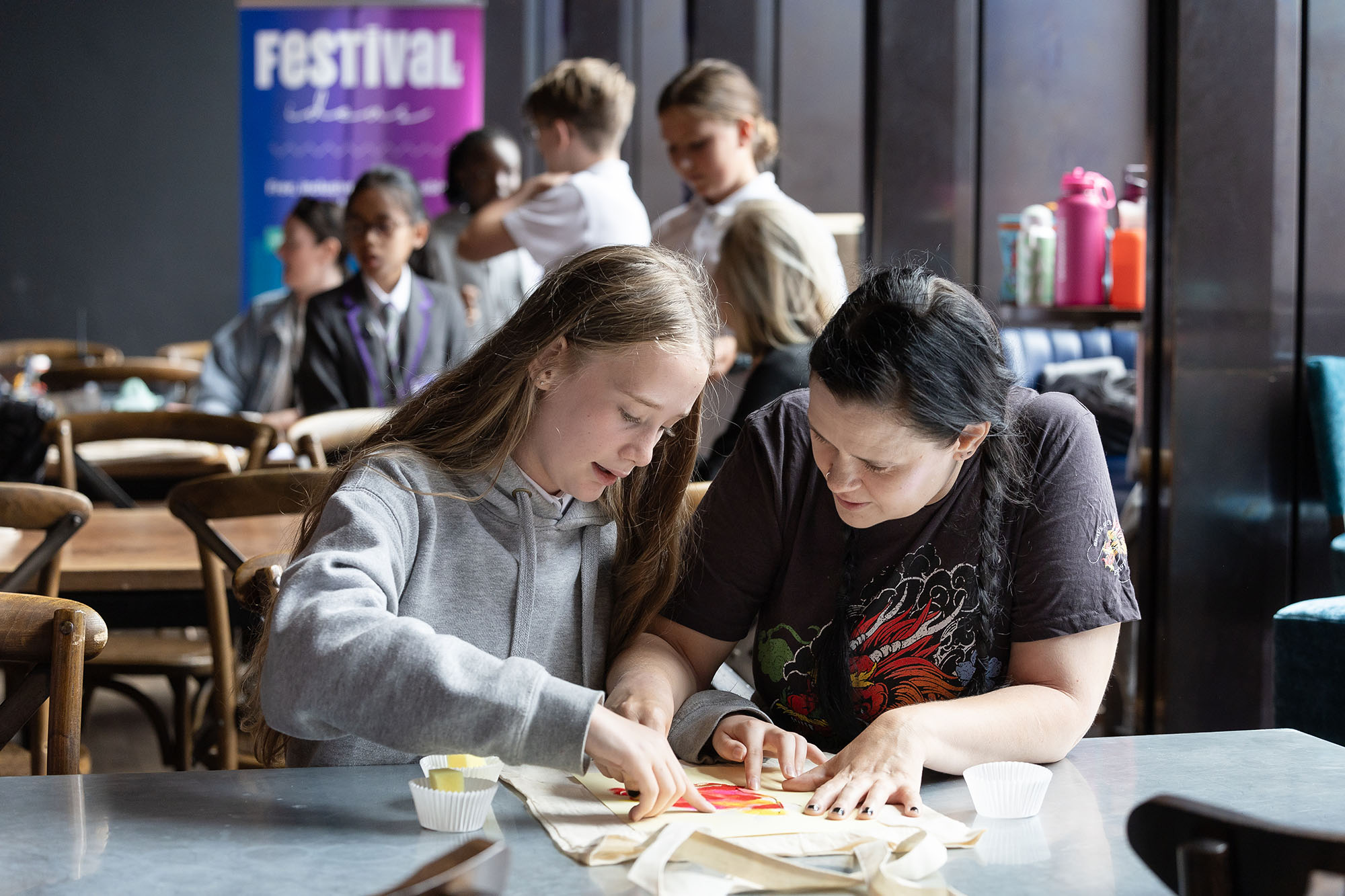 School pupils taking part in a hands‑on arts and crafts workshop at a community festival, creating artwork together at a table with paint and materials, with other students and a Chester Festival of Ideas sign visible in the background.