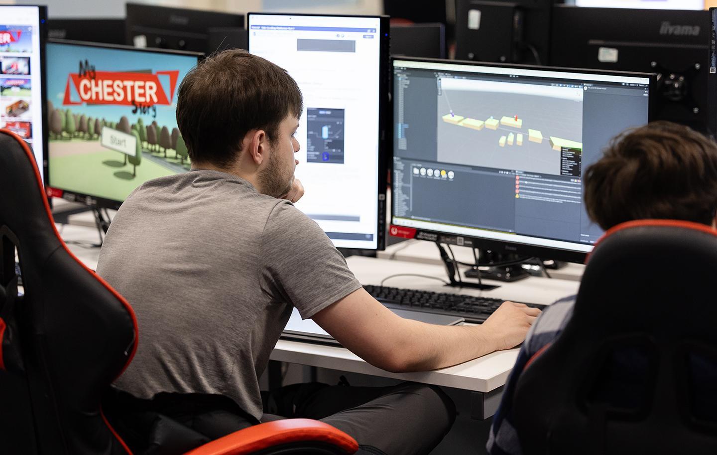 Two men seated at a desk with multiple computer screens, showcasing teamwork in a game design BSc course setting.