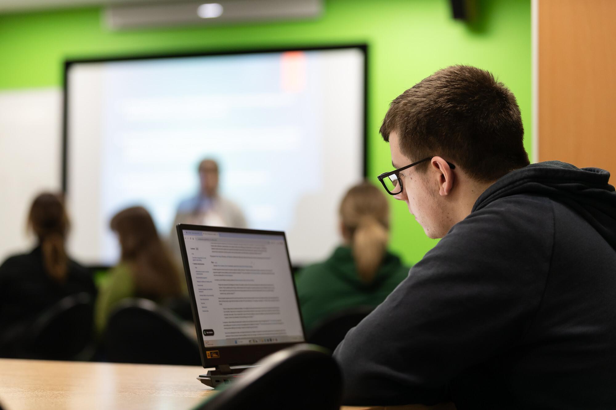 A Criminology with Policing BSc Hons student takes notes on a laptop during a criminology lecture, with a lecturer presenting in the background.