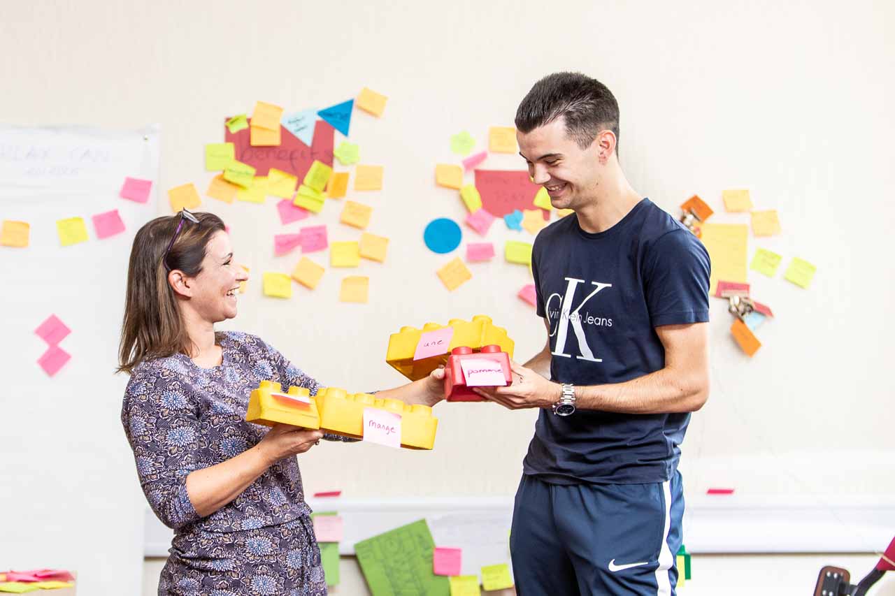 A female tutor smiling and talking to a tall male student holding teaching materials.
