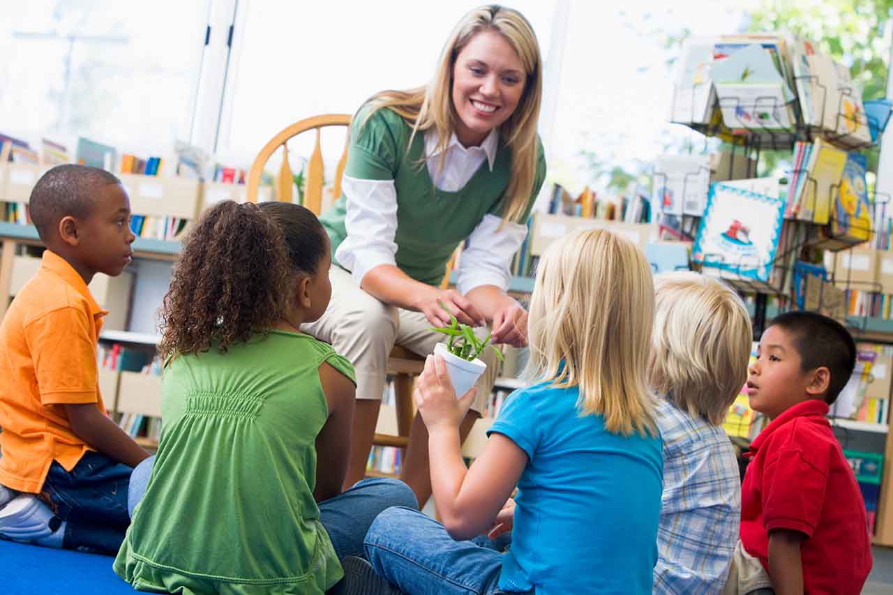 A group of children sitting in classroom with teacher, one child is holding a small green plant in a white pot.