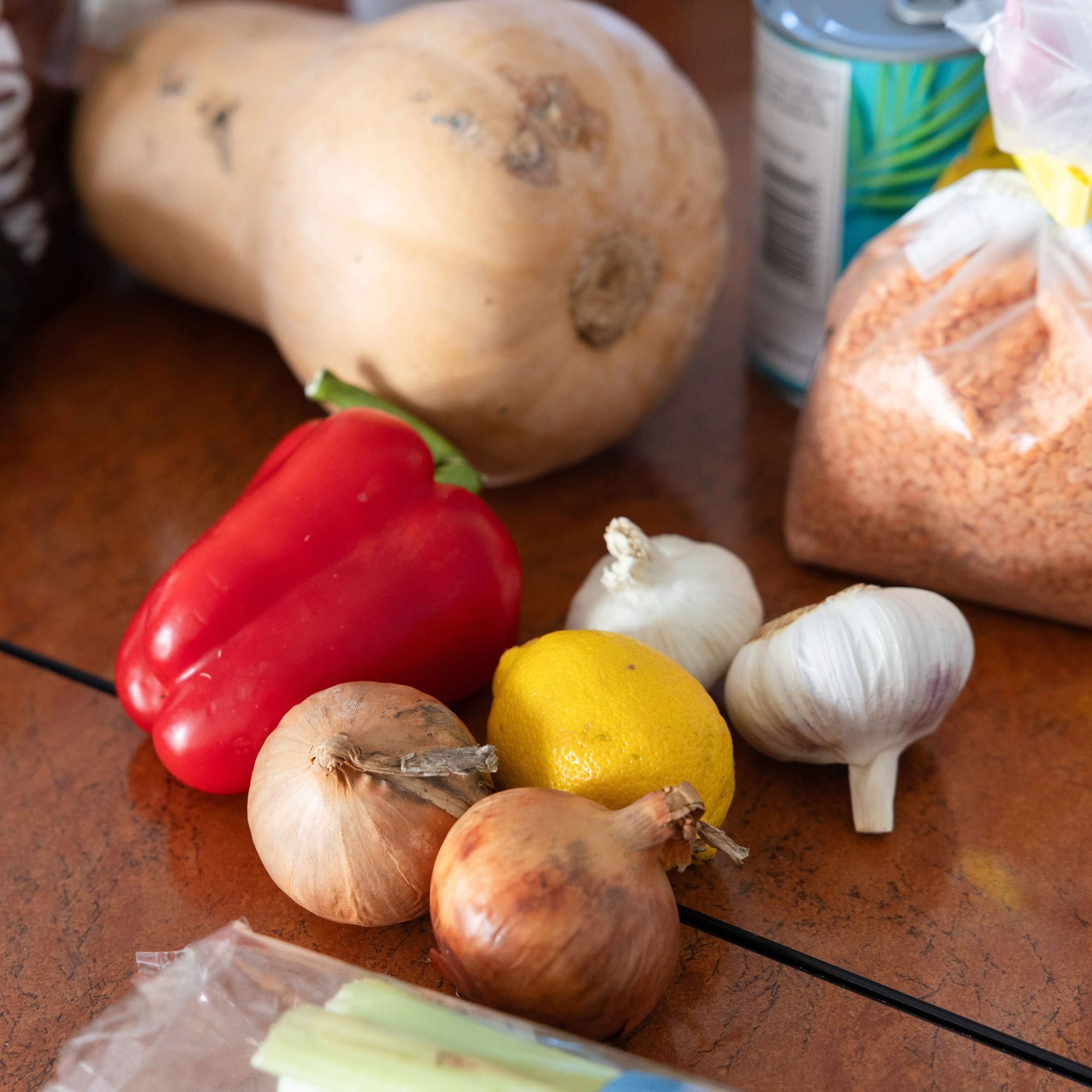 Fresh ingredients on a table, including a butternut squash, red pepper, onions, garlic, lemon and a bag of red lentils.