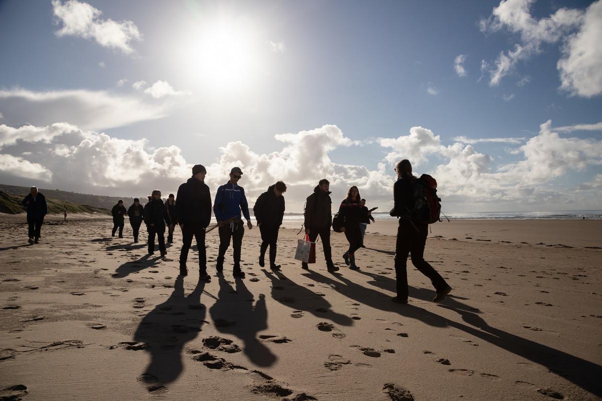 Geography students on a coastal field trip, walking together along a sandy beach under a bright sky.