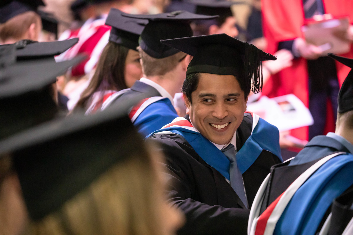 A graduate in a cluter of other graduates sitting down during the ceremony looks over his shoulder to smile at the row behind.