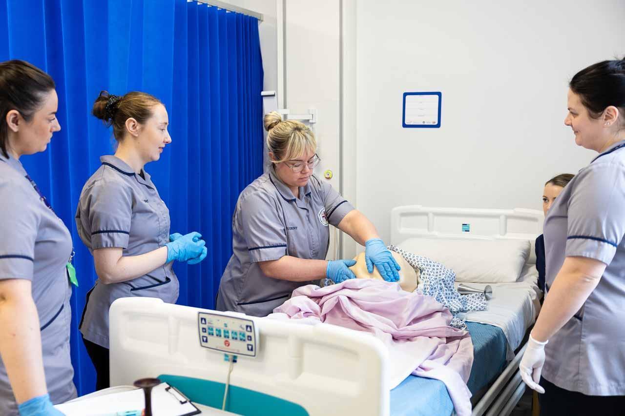 Midwifery BSc degree students in a clinical skills lab practicing maternity care techniques on a training mannequin.