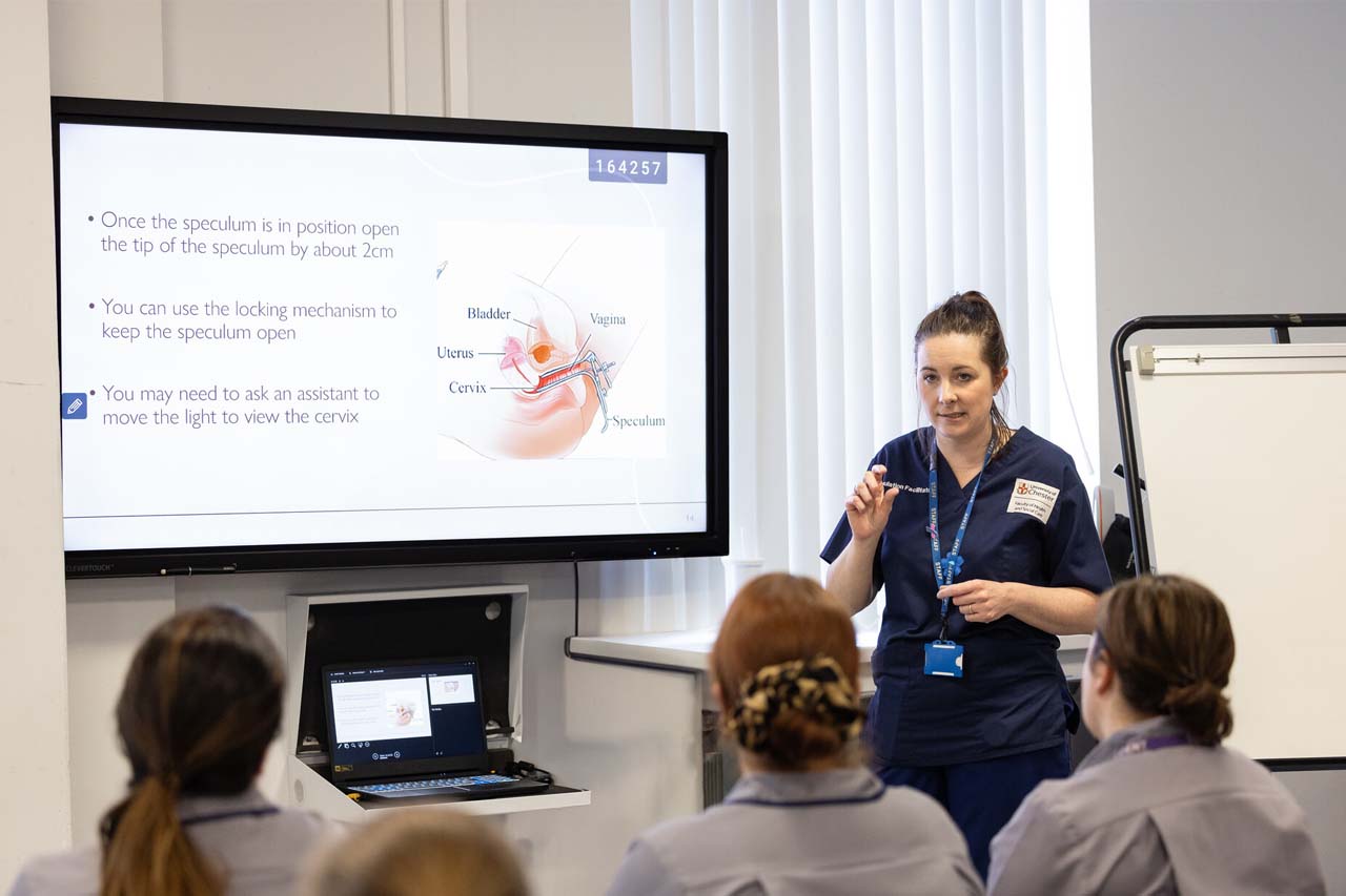 A nurse tutor standing by wall screen, teaching midwifery to group of female nurses.