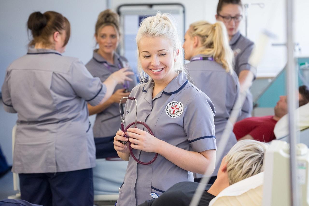 Smiling nursing student in uniform holds a stethoscope while interacting with a patient during a simulated hospital ward session for their Adult and Children's Nursing MNurs Integrated Masters. 