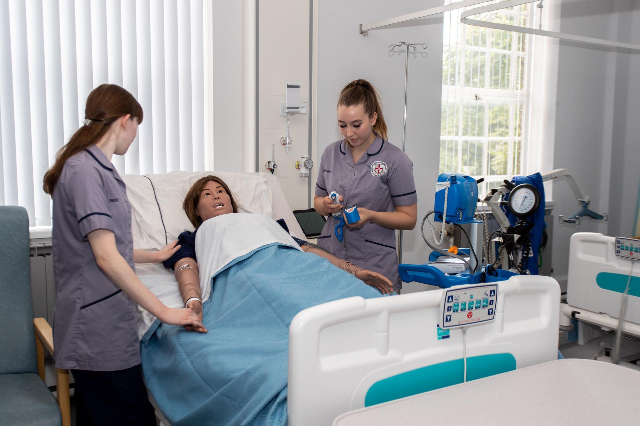 Adult Nursing MSc students practising clinical skills in a simulated hospital ward, providing bedside care to a patient and using monitoring equipment during a hands‑on training session.