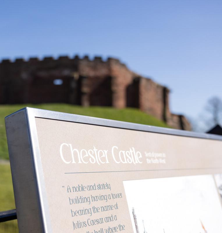 Chester Castle in Cheshire viewed from ground level, with historic stone fortress walls rising behind an informational sign, illustrating a real-world study site for History BA students exploring medieval English heritage and archaeological landscapes.