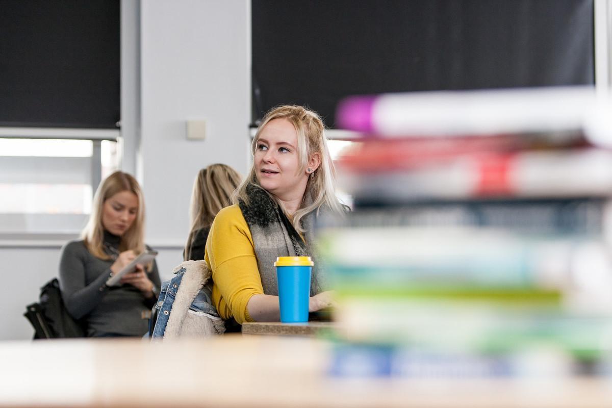 A university student in a classroom listening attentively during a Psychology with Spanish BSc (Hons) session, with books and study materials in the foreground and classmates taking notes in the background.