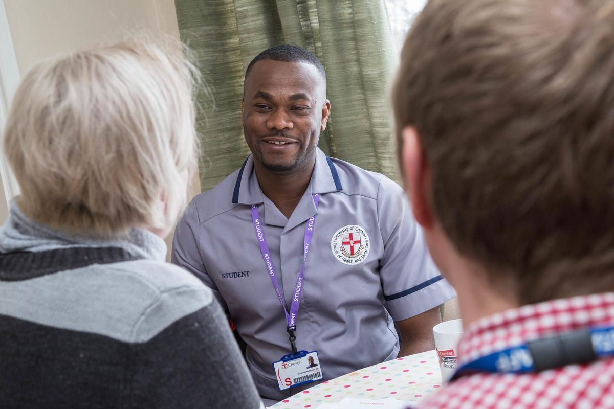  Learning Disability Nursing BN Hons student in uniform, wearing a university ID badge, smiles while talking with two people during a community-based placement, highlighting compassionate care and communication skills.
