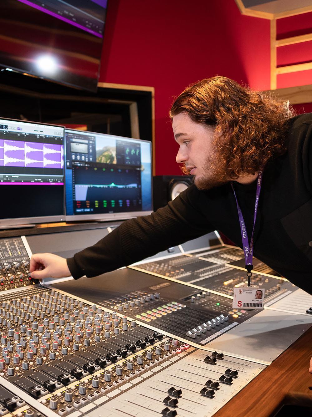 Music Production BA (Hons) student working on a professional mixing console in a recording studio, adjusting sound levels and editing audio on computer software.