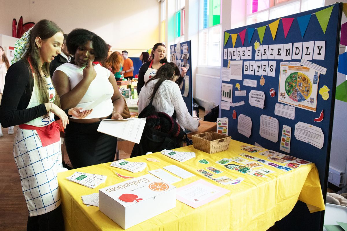 Interest is shown by a student in the 'Budget Friendly Bites' stall at last year's nutrition fair. A number of flyers are on a yellow tablecloth. There are people in the background at other stalls.