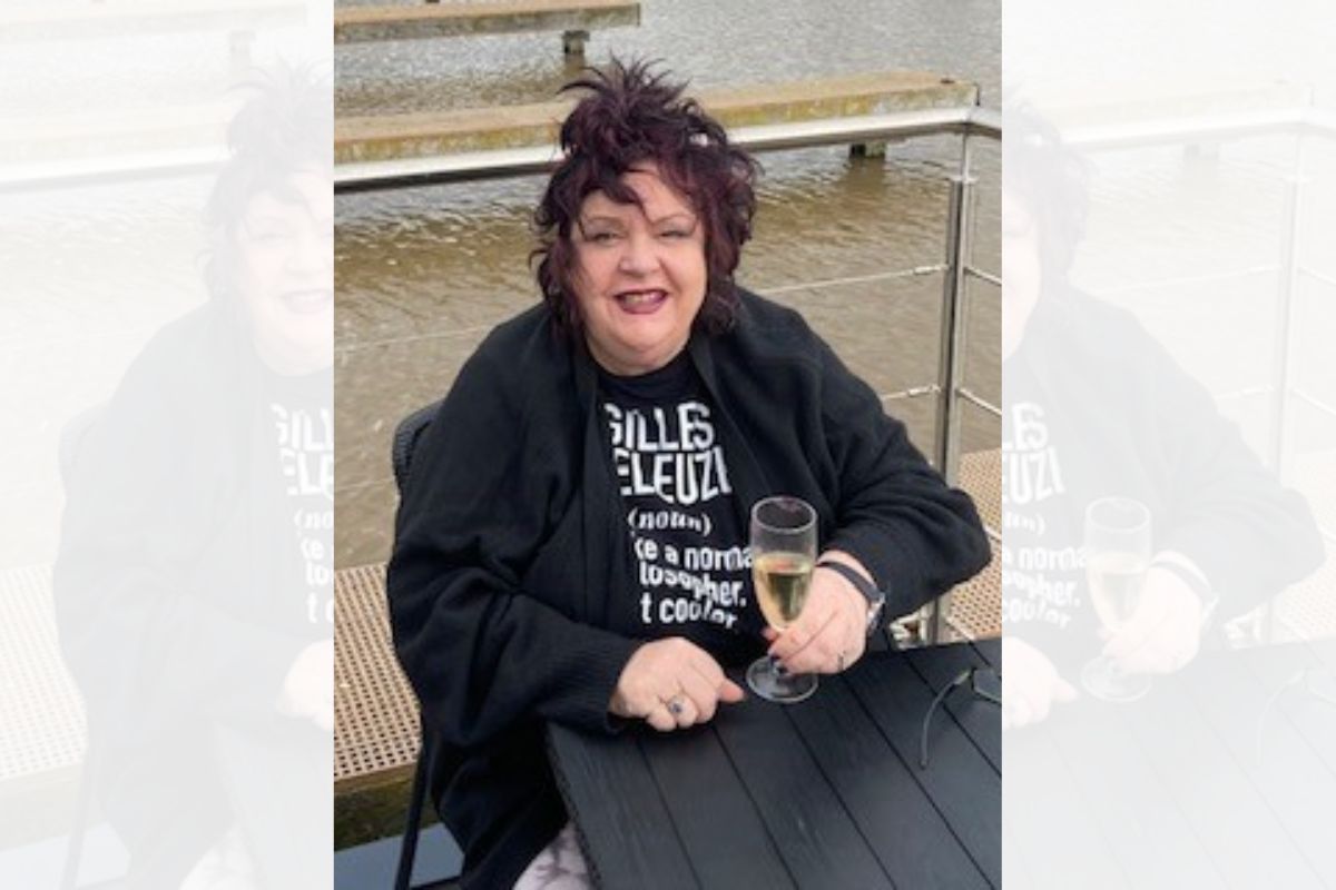 A woman sitting down at a table with a glass of wine looking at the camera. Behind her is a waterway.