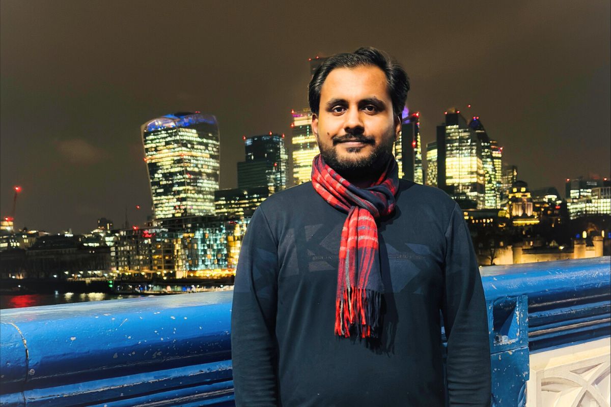 A man wearing a scarf looking at the camera, with a night-time city skyline in the background.