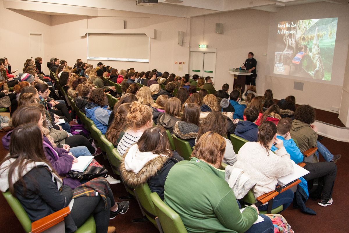 A packed lecture theatre filled with people looking away from the camera towards a lecturer giving a presentation. On a screen in a picture of a child looking at a crocodile, with the Chester Zoo logo in the corner. The title is 'Using the Zoo as a Resource'.
