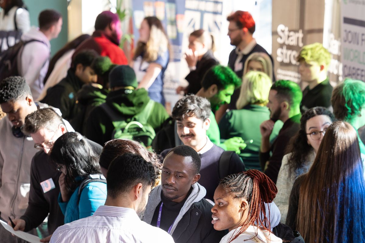A group of people in a busy room looking at stalls and in discussions with people sat at tables.
