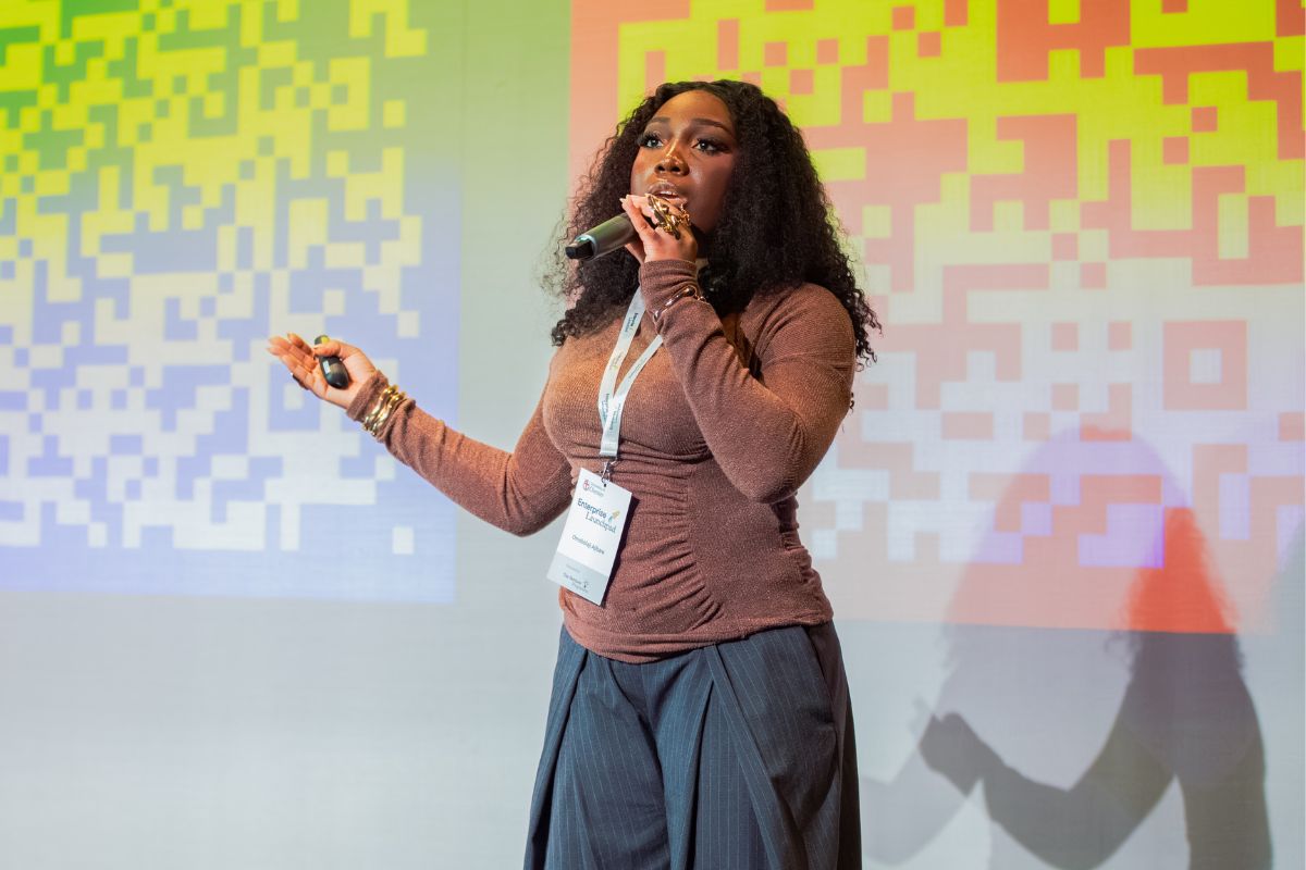 A woman standing, wearing a lanyard, giving a talk to an off-camera audience while holding a microphone to her mouth. In the background are large, colourful QR codes.