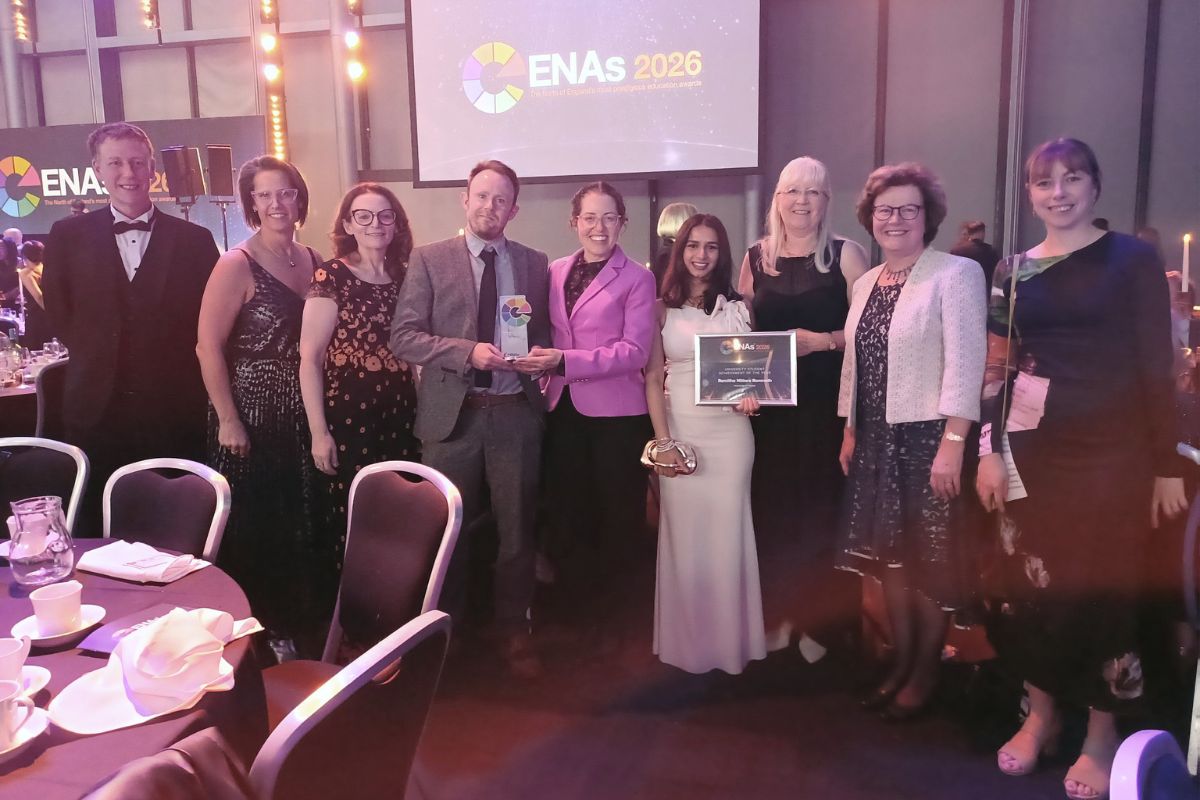 A row of people standing and smiling at the camera in a warmly lit large room. Two of the people are holding an award and another is holding a certificate. The background features the ENAs 2026 logo.