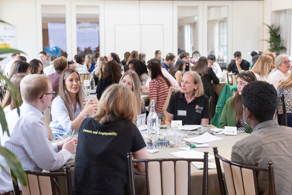 A busy group of people sat at round tables in discussions in a brightly lit room. One of the people as a T-shirt with the words 'Careers and Employability' written on it.