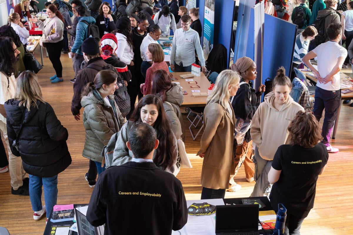 A busy room of people walking around and talking in groups. At the front is a table where two people are stood with Careers and Employability written on their clothing.