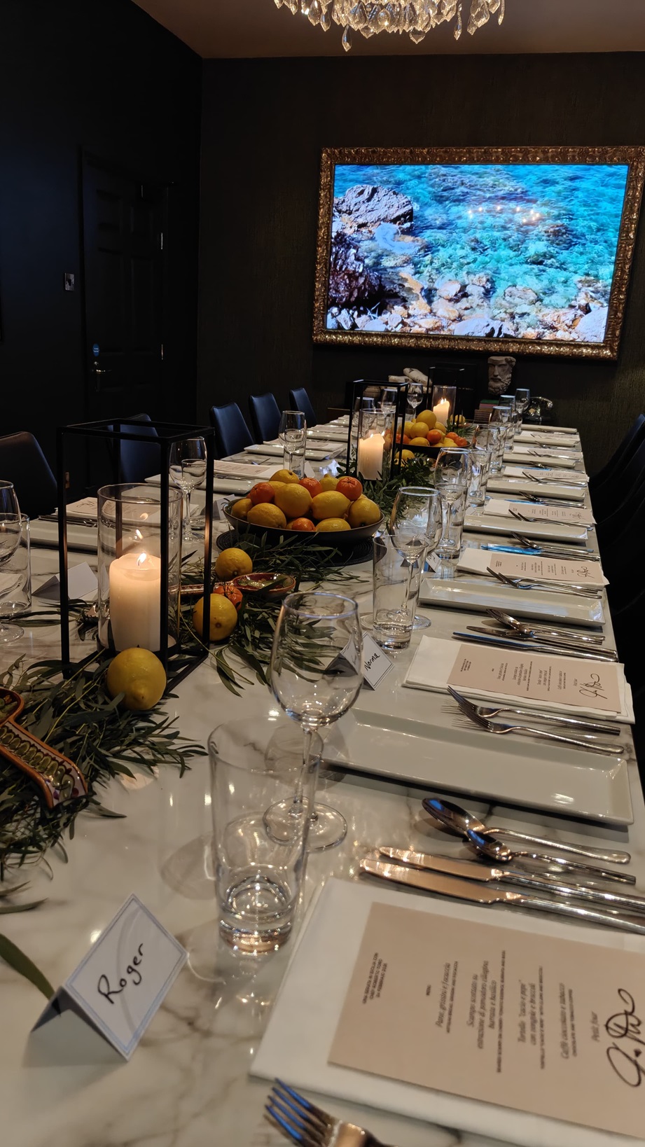 A long table presented for a meal, with decorations, menus, plates, glasses and cutlery. In the background is a framed picture of a rocky shore.