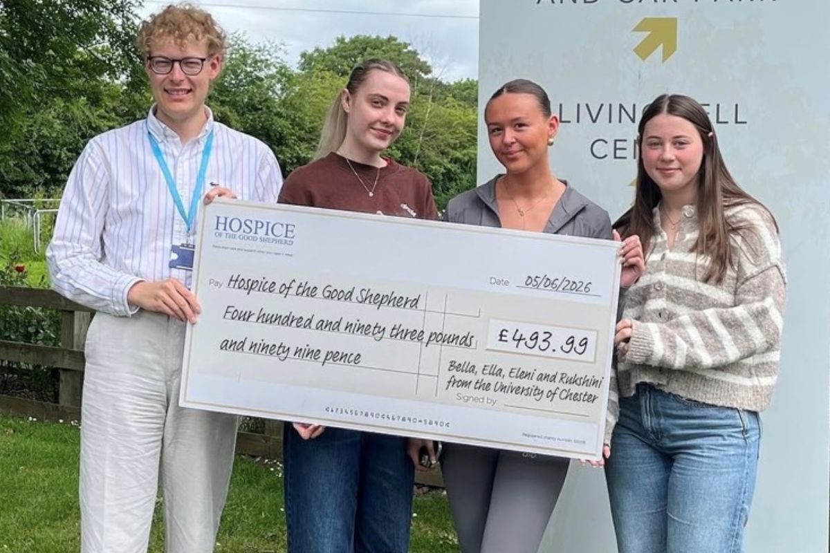 Three students hand over a giant presentation cheque for £493.99 to a representative of the Hospice of the Good Shepherd. There is a sign in the background for the hospice, along with grass, fencing, trees and shrubbery.