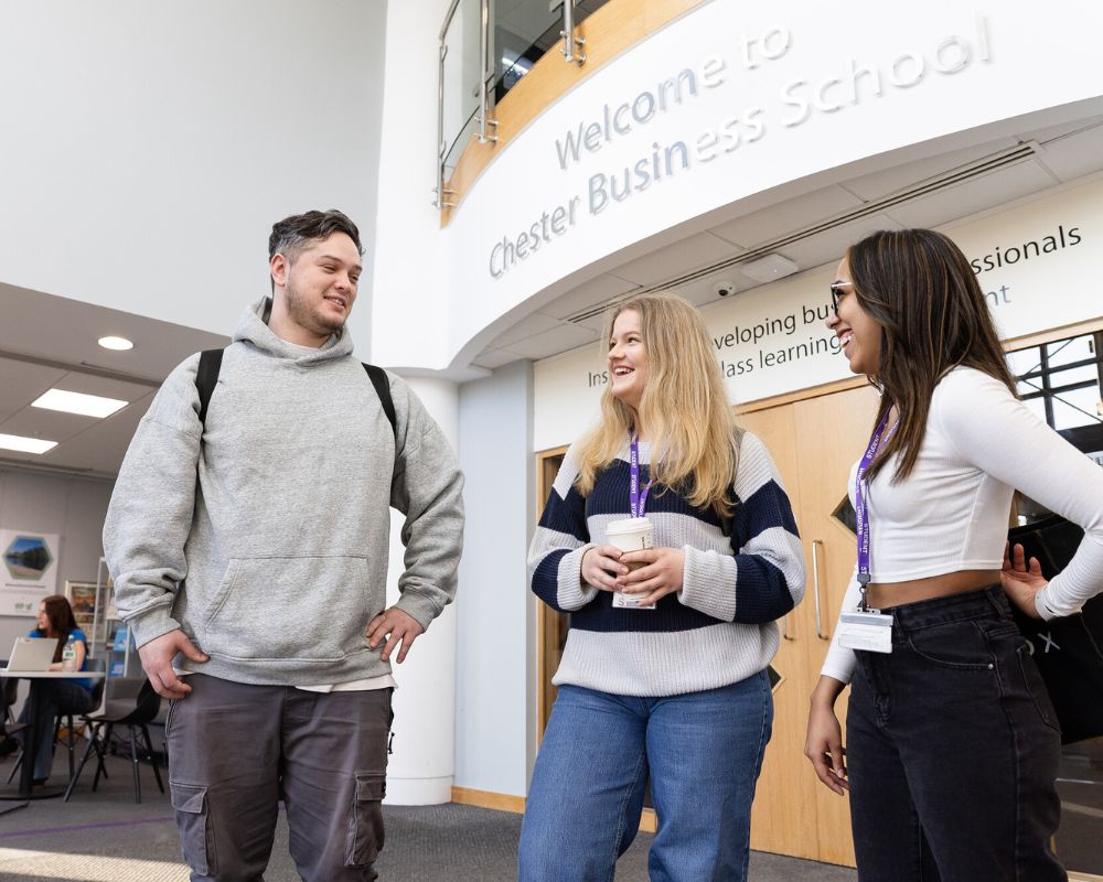 Three people standing in conversation, smiling. In the background is a square archway with the signage 'Welcome to Chester Business School'.
