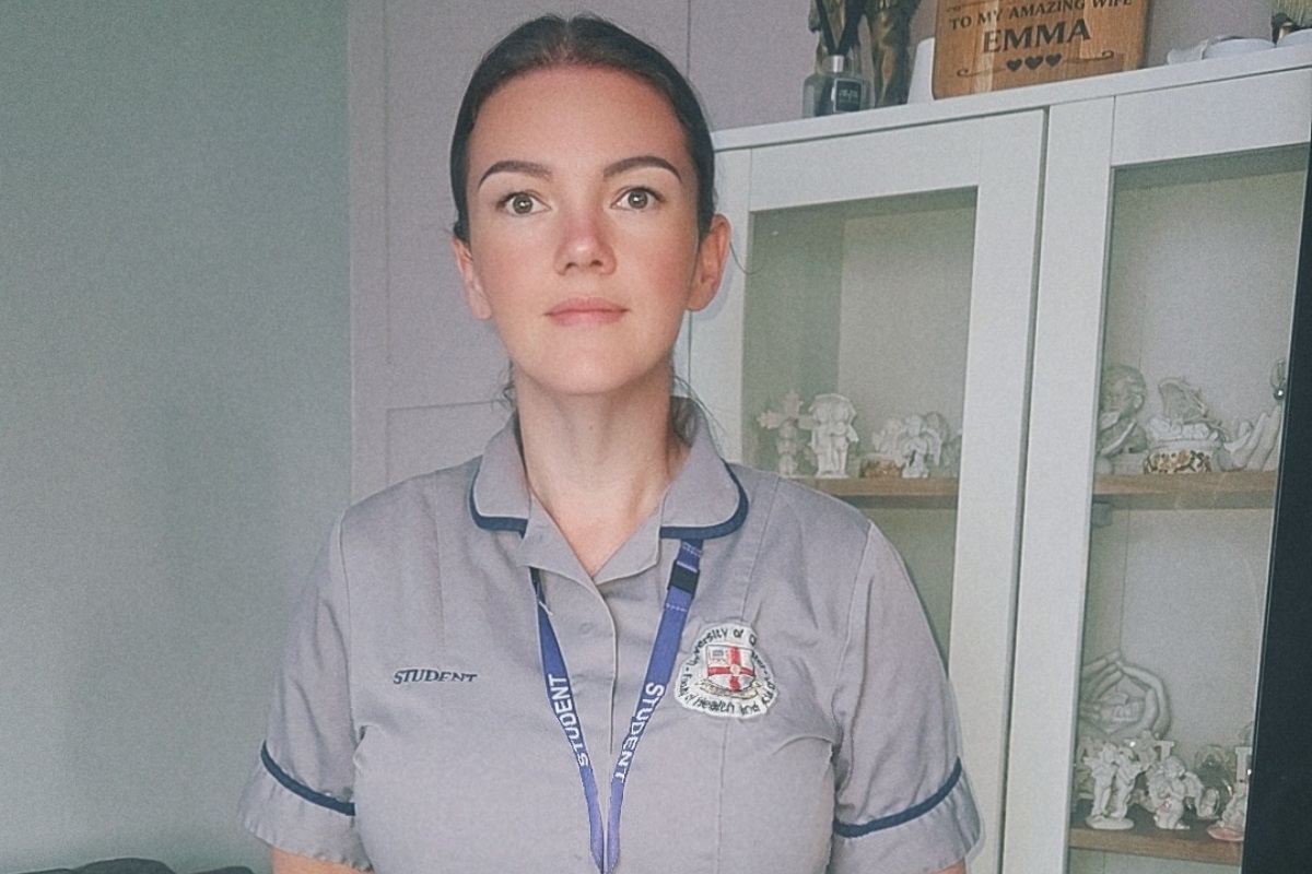 A woman in nursing uniform and a lanyard with 'student' written on it, standing and looking at the camera. In te background is a cabinet wcontaining several ornaments.
