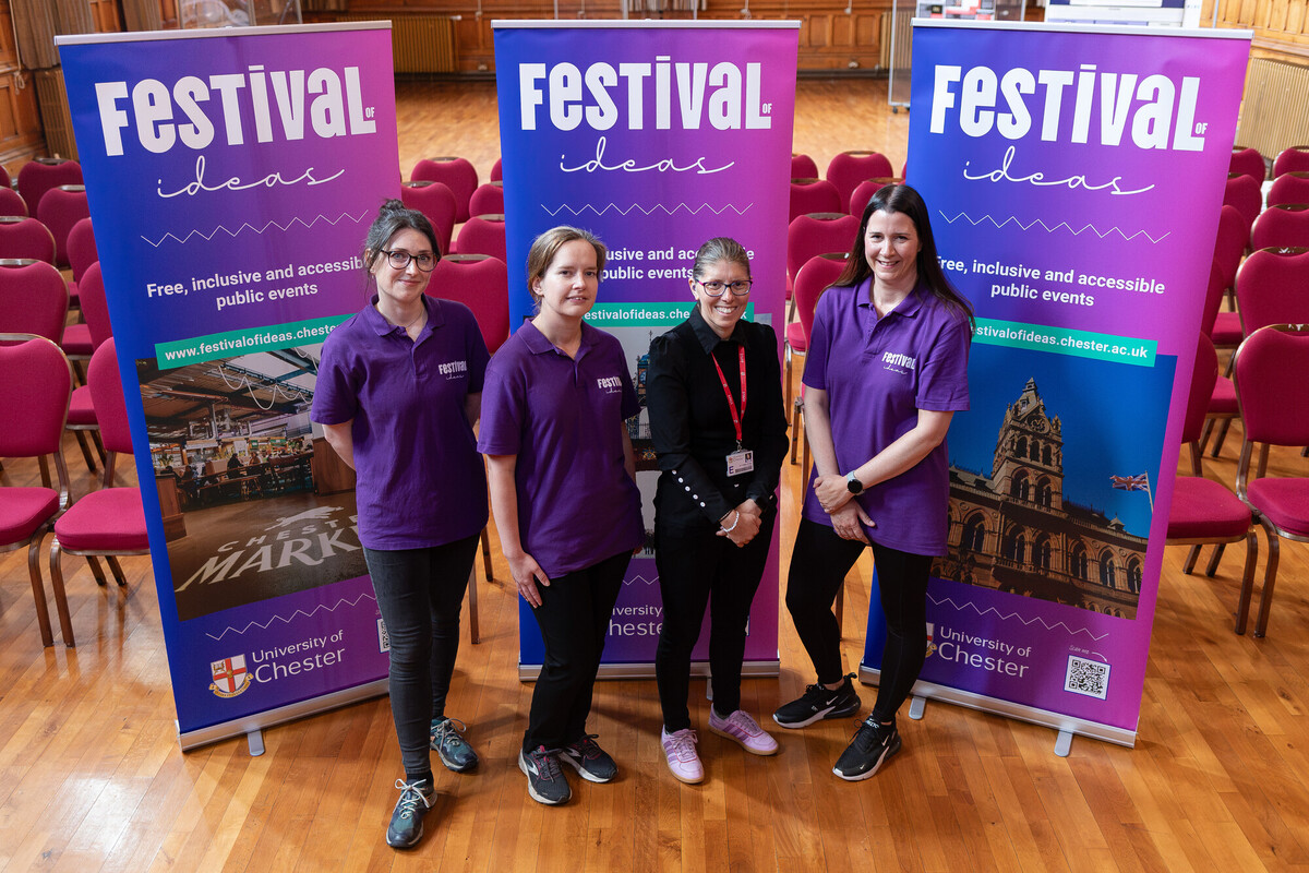 Four women, including Jane Pearson, left, in a row standing and smiling at the camera. In the background are large Festival of Ideas banners.