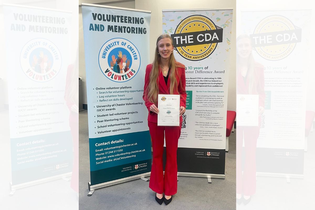 A young woman standing and smiling at the camera, holding a certificate, in front of two tall banners promoting University of Chester volunteers and The Chester Difference Award (The CDA).