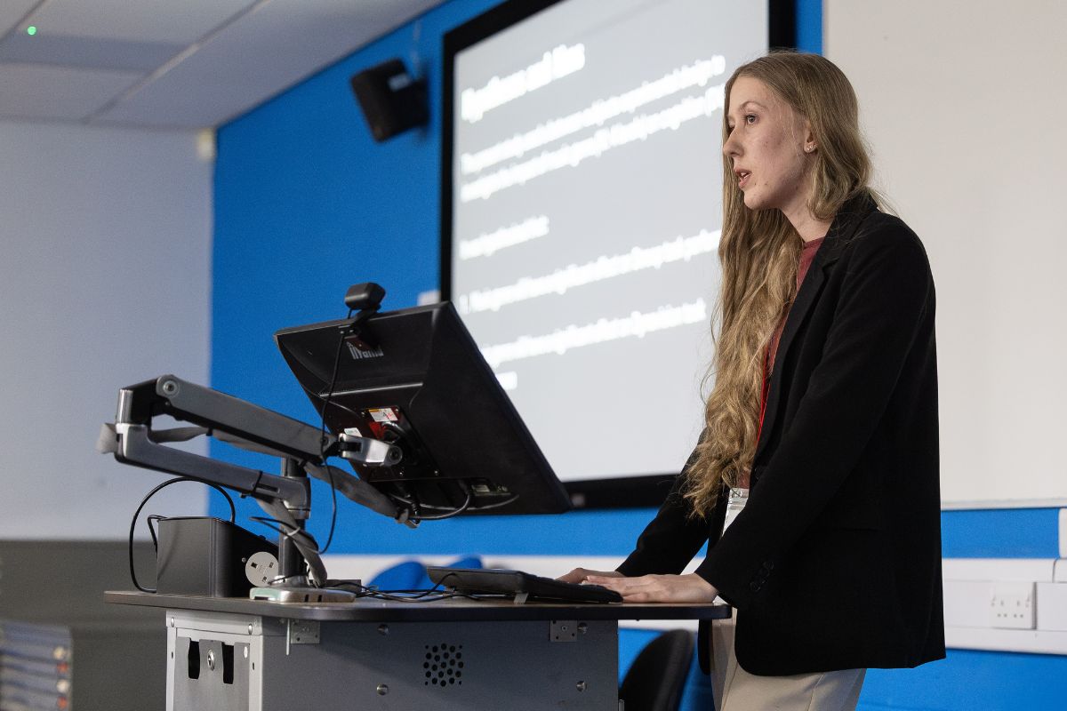 A woman with long hair giving a presentation standing at a desk by a computer monitor. In the background is a projected screen of text which is out of the camera's focus.