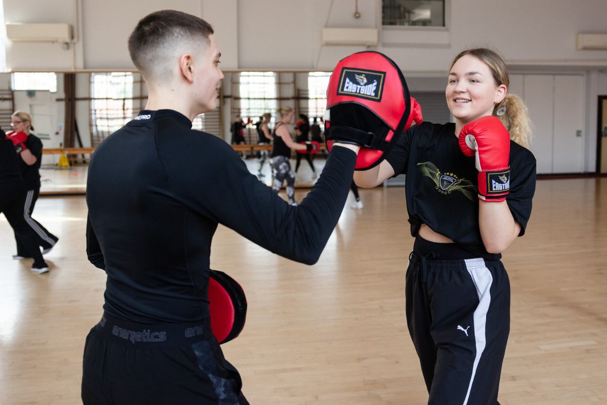 Two students engaged in a boxing training session. The male student, left, is holding a pad for the female student, right, who is punching into it.