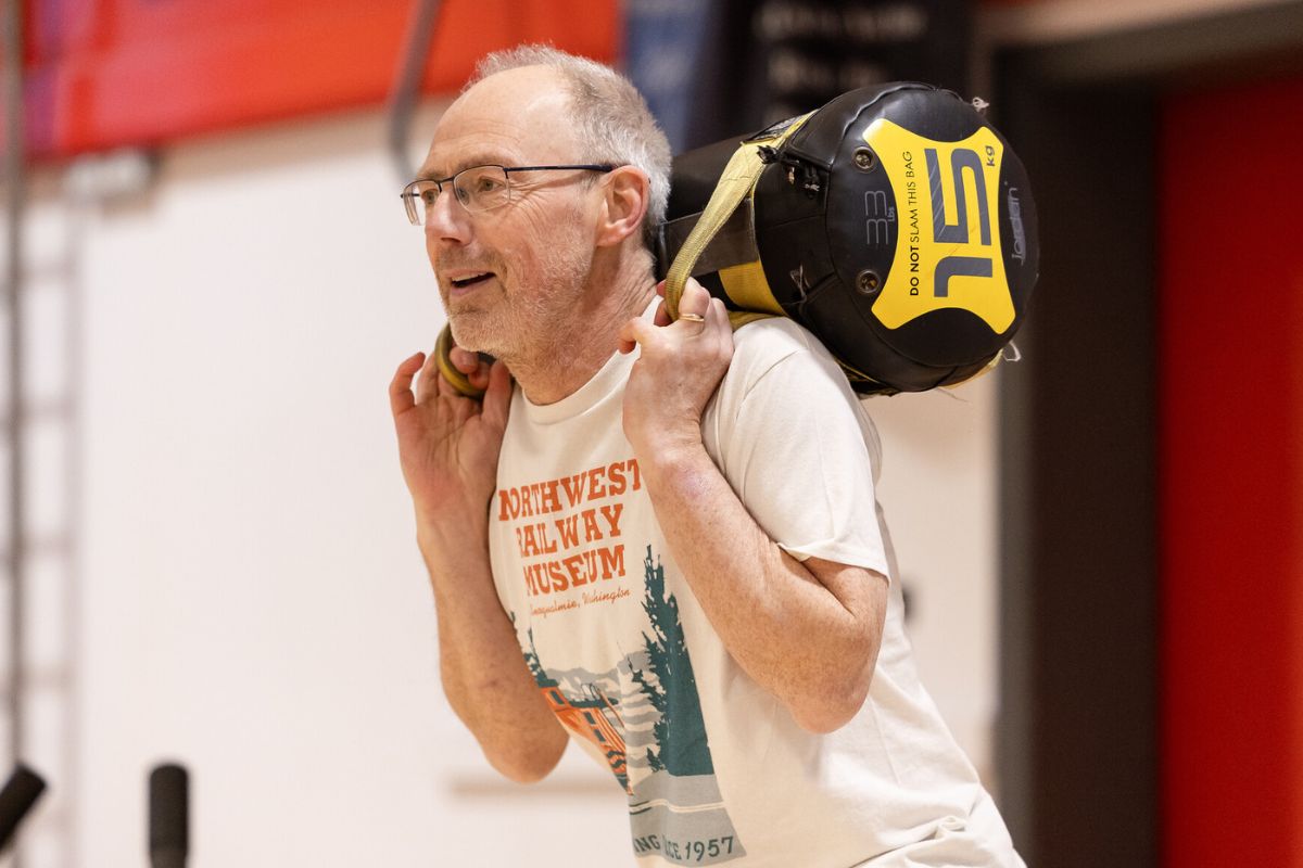 A male member of staff exercising by pulling on straps to keep a weight lifted at shoulder height.