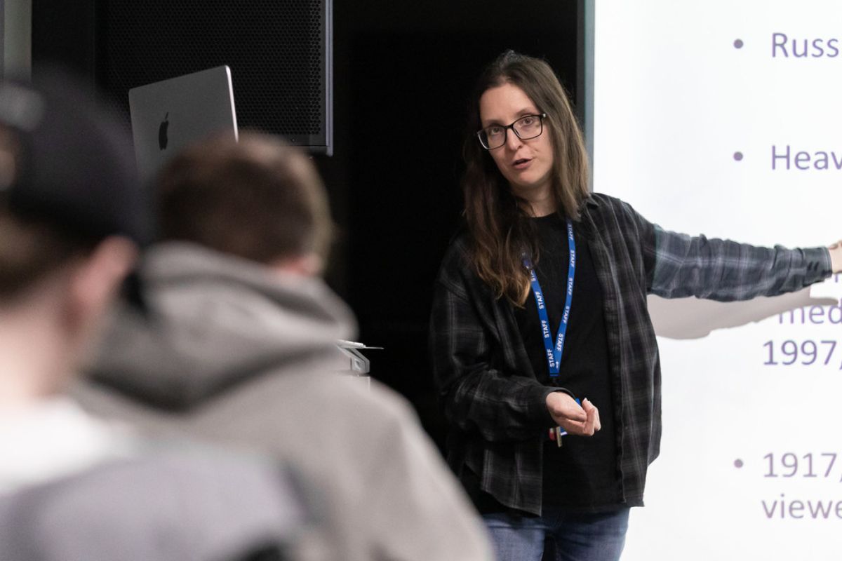 A woman giving a lecture, pointing to a display that's out of the picture, while the backs of two men are at the front of the image, out of focus.
