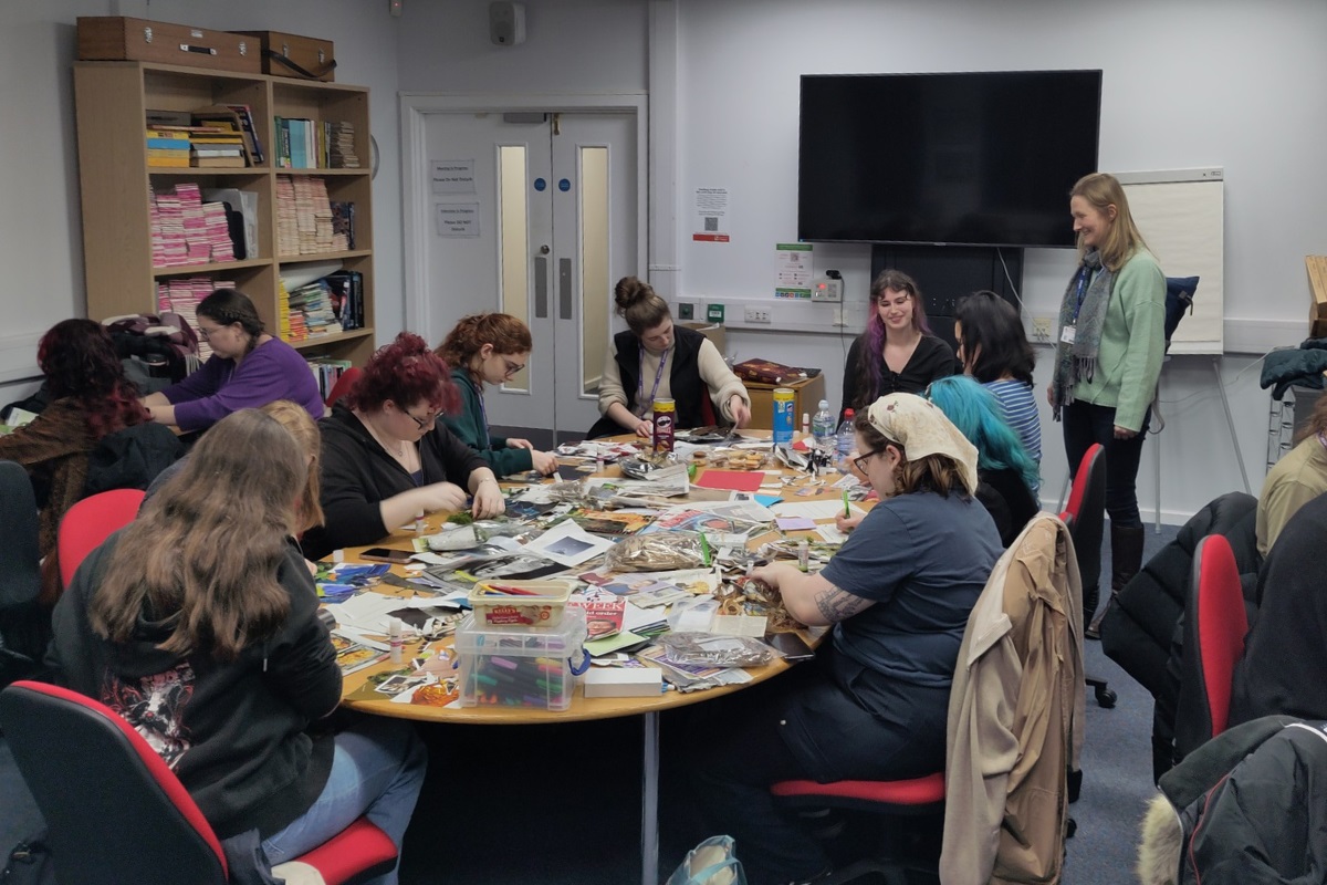 People sat working at a table filled with magazines, arts and craft materials. A lecturer looks on, smiling.