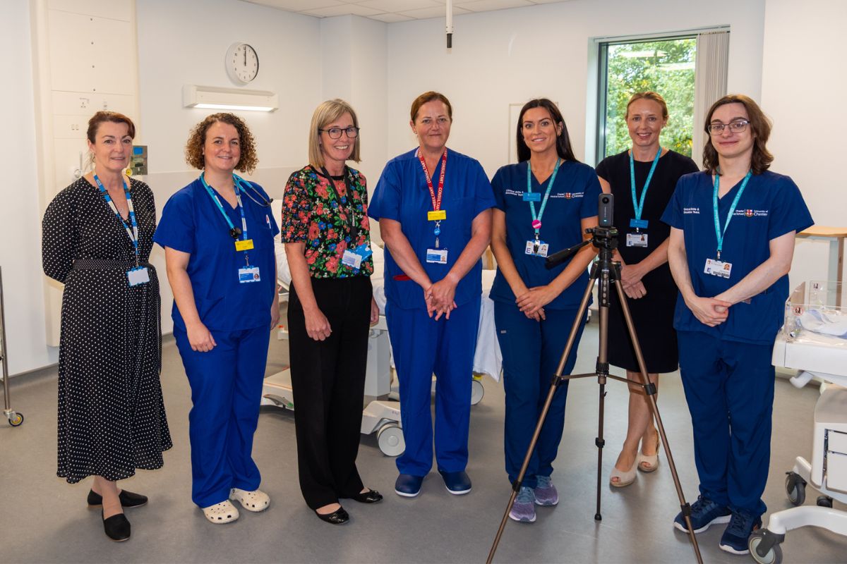 A group of seven people smiling at the camera in a hospital setting. Most of them are in nursing uniforms. There is also a camera mounted on a tripod.
