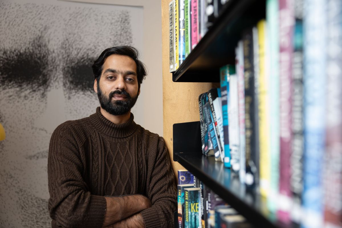 A man standing with his arms folded, looking at the camera, while next to a packed bookshelf.