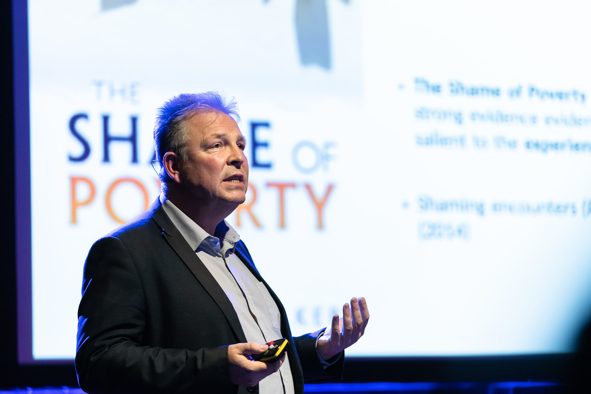 Professor Paul Bissell giving a lecture with a screen behind him. The words are partially obscured but read 'The Shame of Poverty'