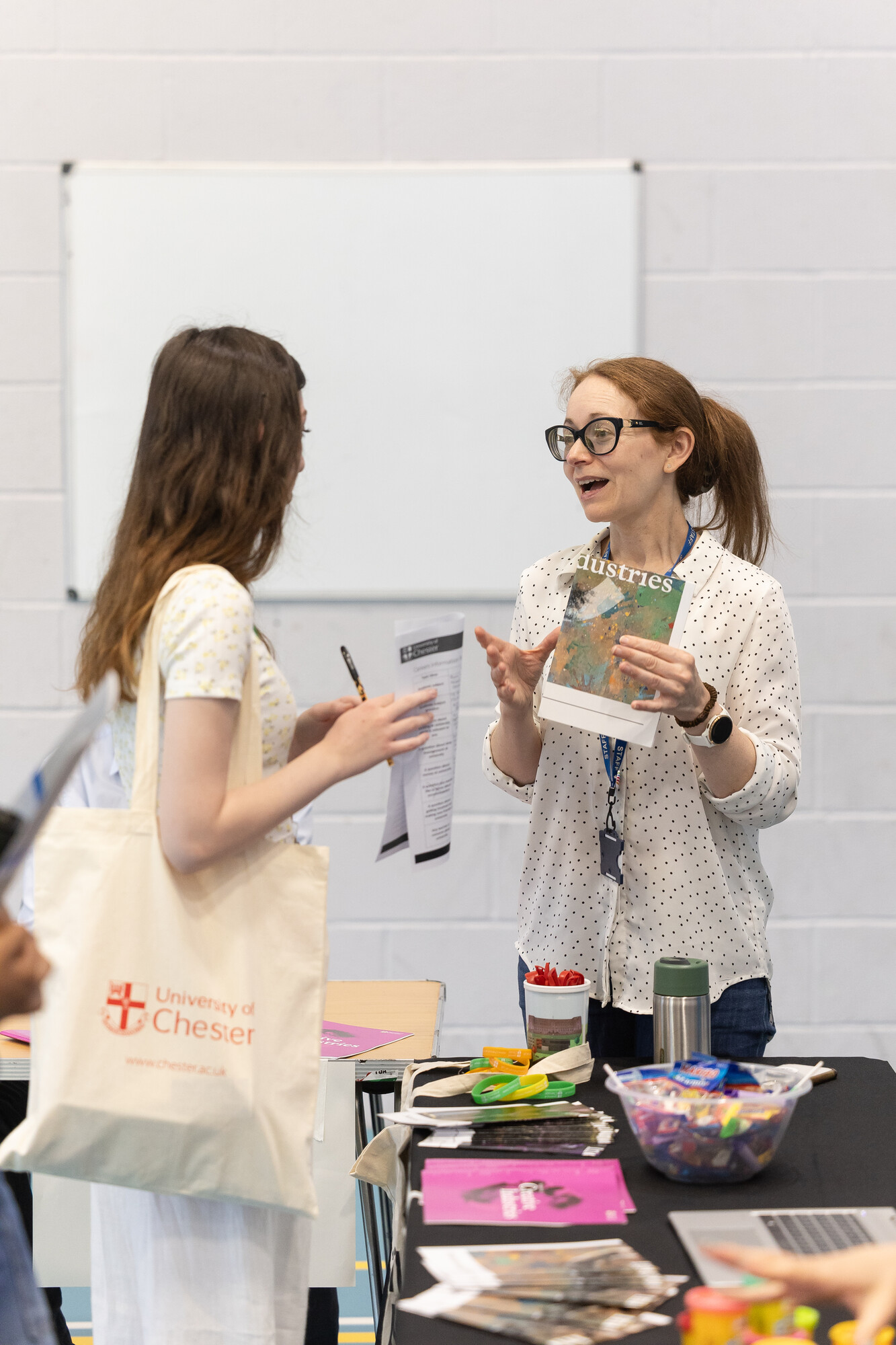 Two females talking to each other. The one on the left is carrying a bag with the University of Chester logo on it. The woman on the right is holding a pamphlet. On the table between them are a variety of small objects including rubber wristbands, pamphlets, and sweets.