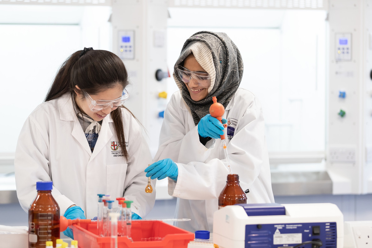 Two students in lab clothing working in a lab; both are smiling as one passes the other a liquid in a tiny container.