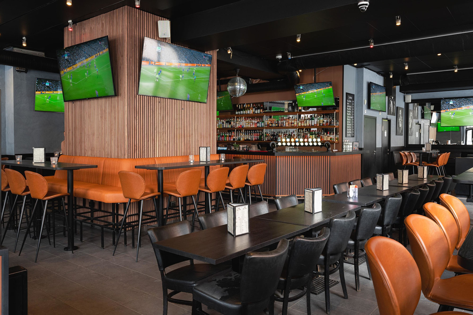 Stylish sports bar with orange seating, black tables, multiple TVs showing football, and a well-stocked bar in the background.