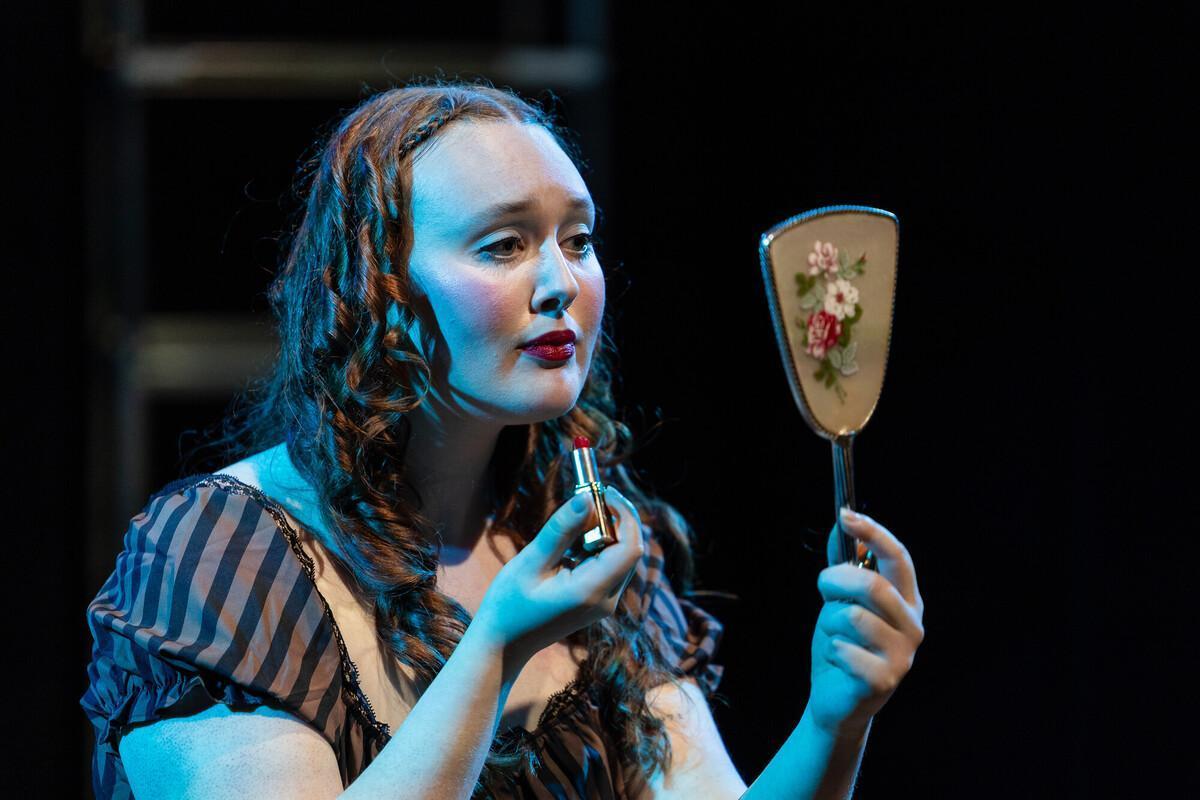 Acting BA student in costume applies red lipstick while looking into a vintage hand mirror on stage, under dramatic lighting, during a theatre for their course. 