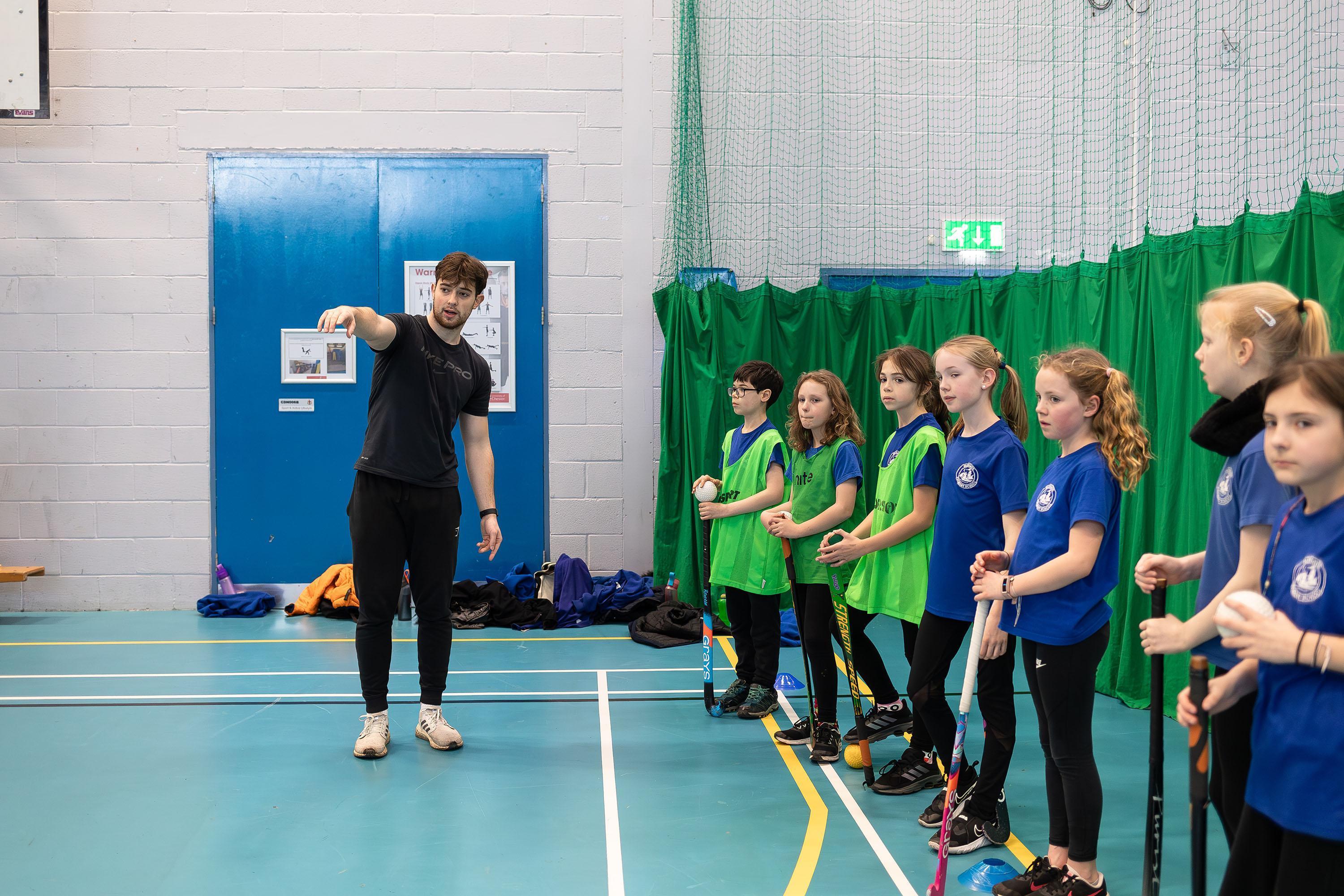University of Chester PE and Sports Coaching BSc Hons student leading a primary school hockey session, developing practical teaching and coaching skills in a sports hall.